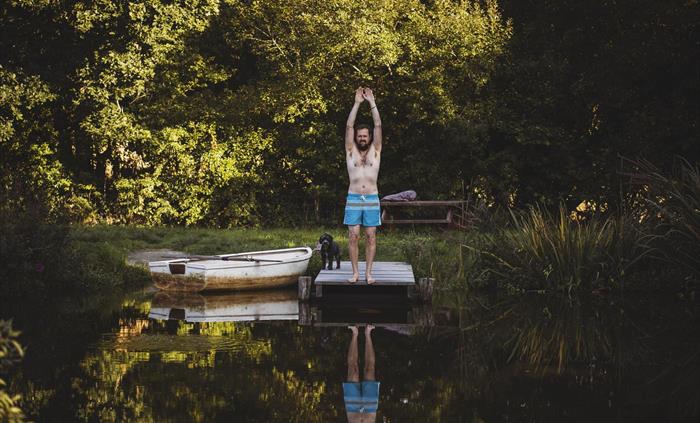 A man standing on a jetty over some water ready to dive. A rowing boat sits to the side of the image. The landscape is woodland.